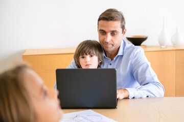 Concentrated father using laptop and holding son. Caucasian middle-aged dad sitting at table with cute little boy and working. Kid looking at camera. Childhood, freelance and fatherhood concept