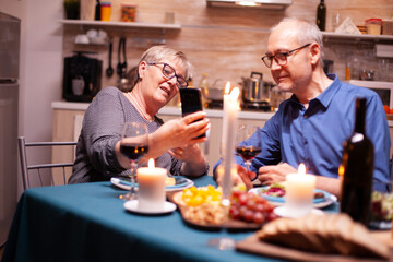 Elderly woman using smartphone to show her husband photos during romantic dinner in kitchen. Sitting at the table in the kitchen, browsing, searching, using phone, internet, celebrating their