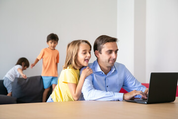 Fototapeta premium Exited lovely girl looking at laptop screen and hugging dad. Caucasian middle-aged father working at home when cute children playing on sofa. Childhood, fatherhood and digital technology concept