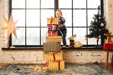 A little boy puts a gift on the top of a big stack of Christmas presents. A panoramic window and a Christmas tree on the background. New Year's eve and Christmas concept.