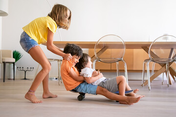 Cheerful children playing with skateboard at home. Blonde adorable girl pushing her two playful brothers. Happy kids riding on board and having fun. Childhood, game activity and weekend concept