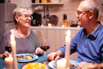 Elderly couple talking about happiness during festive dinner. Happy cheerful senior elderly couple dining together in the cozy kitchen, enjoying the meal, celebrating their anniversary.