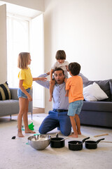 Exited father having fun with children in living room. Happy dad holding son on shoulders. Adorable girl and boy standing near them. Pans and bowl for game. Childhood, weekend and home concept