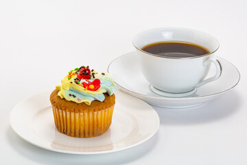 Obraz na płótnie Canvas Vanilla cup cake garnished with butter cream frosting and colorful sprinkles in white ceramic plate and black coffee in white cup on white background with clipping path.