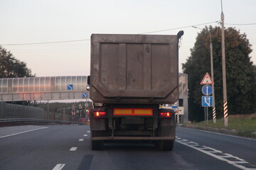 Gray heavy dump truck drive on empty suburban asphalted highway road at summer evening on bridge background rear side view &ndash; Logistics, bulk cargo transportation trucking