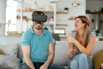 Smiling young man using VR headset at home on couch. Woman and her husband enjoying virtual reality in apartment.