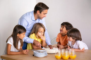 Caucasian father cutting delicious cake for children. Cute little kids surrounding table, celebrating birthday together, talking and waiting for dessert. Childhood, celebration and holiday concept
