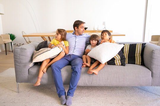 Caucasian dad sitting on sofa and embracing cute kids. Loving middle-aged father relaxing with adorable children on coach in living room and talking. Childhood, family time and fatherhood concept