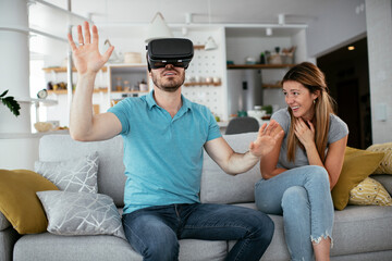  Smiling young man using VR headset at home on couch. Woman and her husband enjoying virtual reality in apartment.