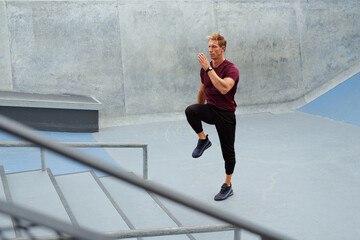 Fototapeta na wymiar Young Man Running On Place Near Concrete Steps Outdoors. Handsome Caucasian Sportsman With Strong Muscular Body In Fashion Sportswear Warming Up Before Intense Workout.