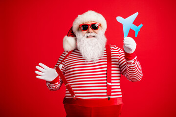 Photo of old man grey beard demonstrate blue paper plane funny wear santa claus x-mas costume suspenders sunglass striped shirt cap isolated red color background