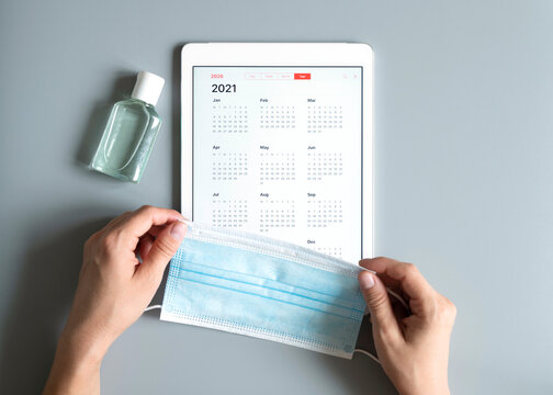 a tablet with an open calendar for 2021 year and protective medical mask and hand sanitizer in woman's hands on a gray background. covid-19 coronavirus protection concept in 2021
