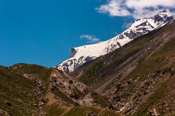 A snow capped mountain peak rising above the green, rugged, slopes of a hill around the Kunzum La pass in the Spiti valley.