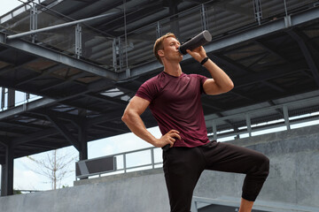 Fototapeta na wymiar Young Man In Sweaty Shirt Drinking From Bottle After Intense Workout Against Concrete Wall. Handsome Caucasian Sportsman With Strong Muscular Body In Fashion Sportswear Having Break.