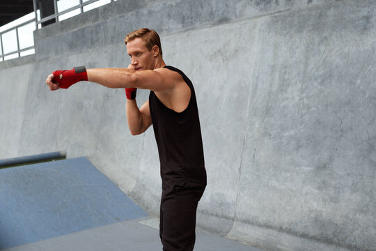 Young Man Stands In Boxing Pose And Doing Punching Workout. Handsome Caucasian Sportsman With Strong Muscular Body In Fashion Sportswear And Hand Wrap On Wrists Against Concrete Wall.