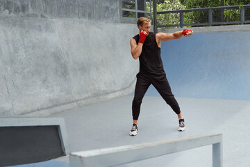 Young Man Stands In Boxing Pose And Doing Punching Workout. Handsome Caucasian Sportsman With Strong Muscular Body In Fashion Sportswear And Hand Wrap On Wrists Against Concrete Wall.