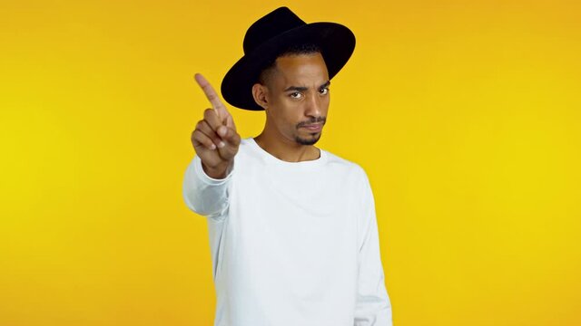 Portrait of african serious man in black hat showing rejecting gesture by stop finger sign. Guy isolated on yellow background.