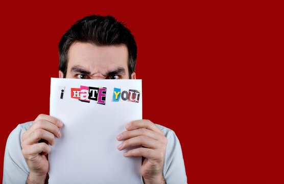 Angrey looking dark haired man holding up paper with the text 'I Hate You'. Letters cut out from paper. Isolated on red background.