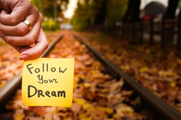 girl holds in her hand a yellow sticker with the inscription follow your dream on the background of an autumn railway or rail