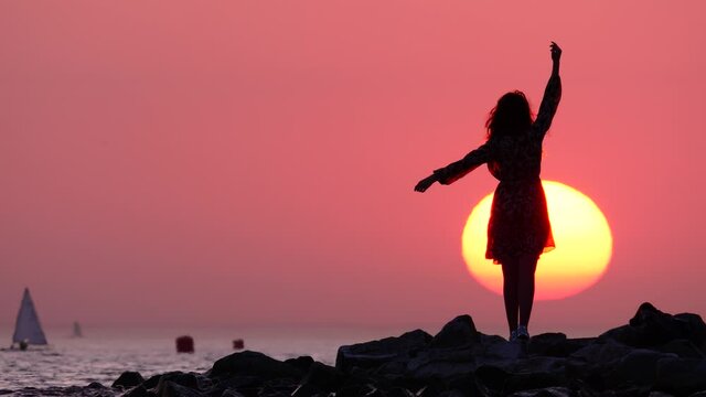 Romantic woman stand on stones at sea shore, she raise and stretch out hand, light dress flutter on wind. Large disk of sunset sun on background, small dinghy sails seen on water