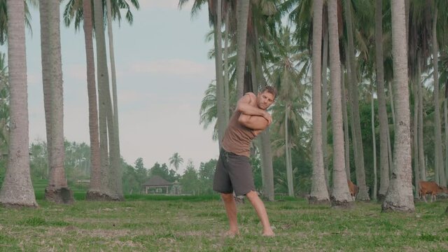 Full Shot Of Middle-aged Sporty Caucasian Man Standing With Hands On Shoulders Rotating Upper Body. Male Fitness Person Warming Up In Front Of Tall Palm Trees And Brown Cows Grazing On Grass