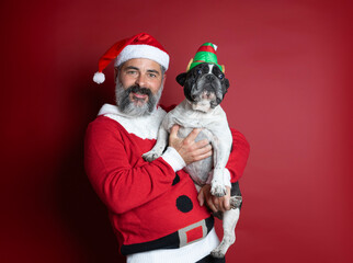 Portrait of man dressed for christmas holding a small french bulldog with elf hat on red...