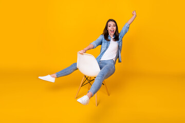 Fototapeta na wymiar Full body photo of attractive pretty funny lady good mood sit chair spread legs fooling around playing raise fist dream riding wear casual denim shirt shoes isolated yellow color background
