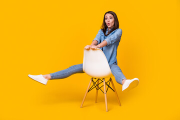 Fototapeta na wymiar Full length photo of attractive pretty funny lady good mood sitting comfy chair spread legs hips fooling around wear casual denim shirt white shoes isolated bright yellow color background