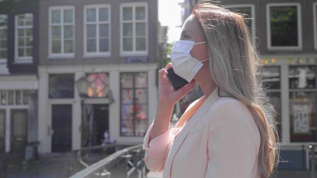A Casual Dutch Woman Having Conversation Using Her Smartphone Wearing White Facemask On A Canal Bridge In Red Light District, Amsterdam. - Covid 19 Pandemic - Medium Shot, Slow Motion