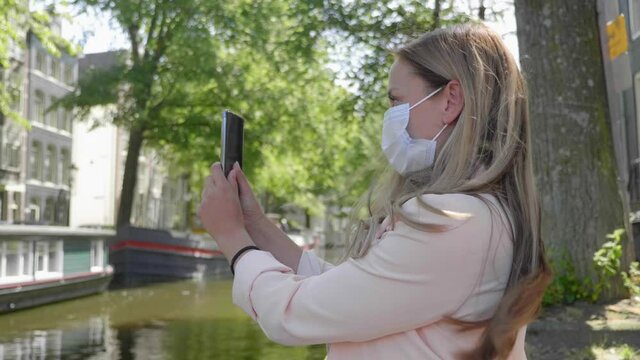 Pretty Woman With Surgical Mask Takes Selfie By The Amsterdam Canal Amidst Covid-19 Lockdown In Netherlands - medium shot