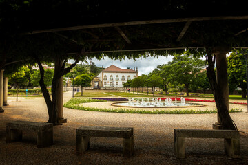City park in Castelo Branco, Portugal, with benches and a shady pergola framing the garden, lake and the old Episcopal Palace in the background, on a spring day with clouds.