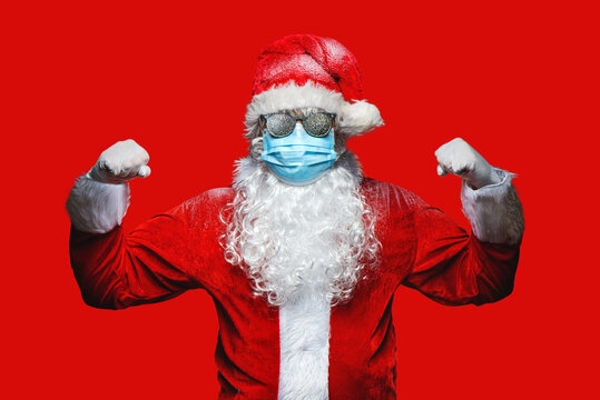 Close up portrait of funny old, cool, bearded  Santa Claus wearing costume, glasses, face mask looking at camera. Covid 19 coronavirus safety protection concept.