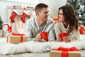 Happy couple with cups of hot drink on floor in room decorated for Christmas