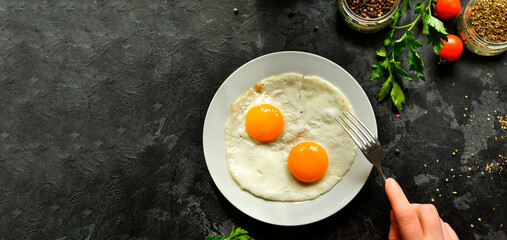 Woman eats fried eggs for breakfast, holds a fork. Fried eggs in a plate. Top view, dark background. Breakfast and ingredients.