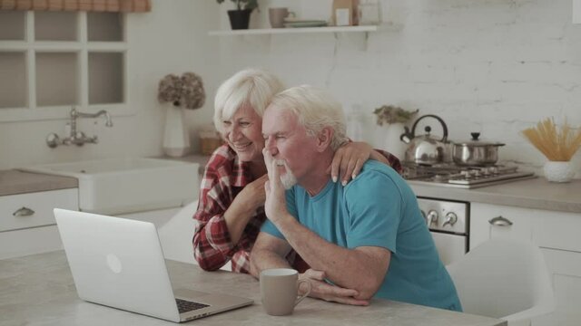 Happy grandparents sitting by table in the kitchen and waving hands to their friends or relatives while talking through video-chat