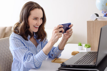 smiling brunette woman with smartphone in hands sitting at laptop, video games, video calls concept