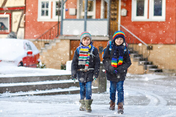 Fototapeta na wymiar Two little kids boys of elementary class walking to school during snowfall. Happy children having fun and playing with first snow. Siblings ans friends with backpack in colorful winter clothes.