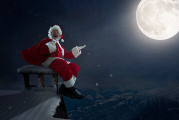 Obraz premium Emotional Santa Claus congratulating with New Year 2021 and Christmas. Man in traditional costume sitting on house roof with full moon on background in midnight. Winter, holidays, sales. Copyspace.
