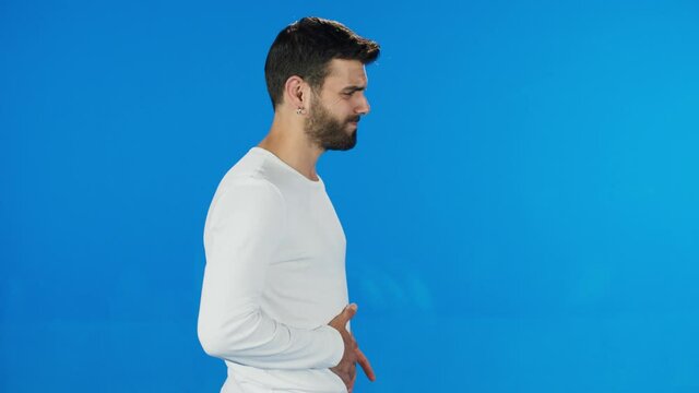 Male suffering from digestive problems. Bearded man wearing white t-shirt sudden stomach ache, severe abdominal pain, constipation, gastritis, pancreatic diseases. Blue background.