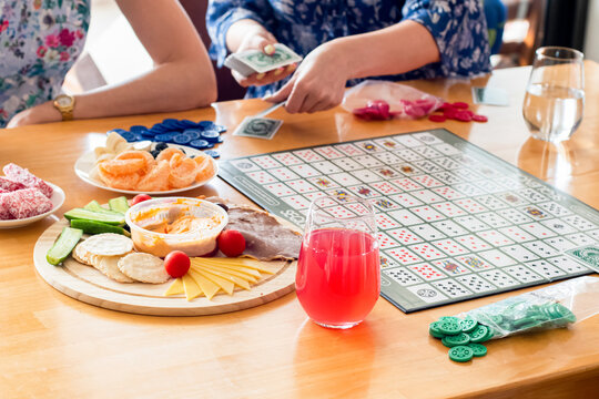 Friends Ready To Play Sequence Board Game With Color Chips And Playing Cards. Hands Dealing Cards To Start The Game. Cheese Platter Board And Drinks On Side.