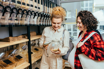 Two female sisters with cotton bag and glass jar buying soy in zero waste shop. Dispensers for cereals, grains, nuts in plastic free grocery store. Sustainable shopping at small local businesses.
