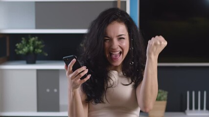 Excited young woman rejoices at message on her smartphone. Happy female looking at cell phone celebrate good news victory success. Lucky gambler showing yes winner gesture.