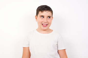 Caucasian young boy standing against white background  with thoughtful expression, looks away keeps hands down bitting his lip thinks about something pleasant.