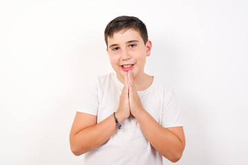 Caucasian young boy standing against white background  keeps palms together, has pleased...