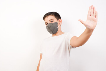 Caucasian young boy wearing medical mask standing against white background doing stop gesture with palm of the hand. Warning expression with negative and serious gesture on the face.