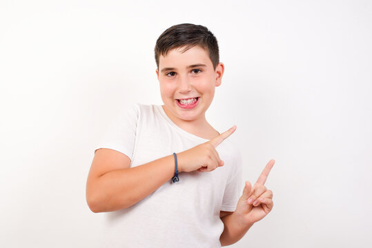 Positive Caucasian young boy standing against white background with beaming smile pointing with two fingers and looking on empty copy space. Advertisement concept.