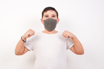 Portrait of Caucasian young boy wearing medical mask standing against white background  looks with excitement at camera, keeps hands raised over head, notices something reacts on sudden news.