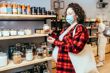 Pregnant woman with a cotton shopping bag wearing a protective face mask choosing foods in a plastic free grocery store. Sustainable shopping at small local businesses.