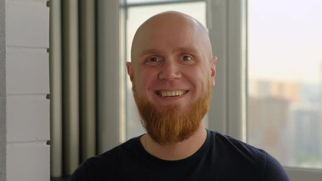 A young, Caucasian, bald guy with a red beard sits by the window, looks into the camera and smiles a pleasant friendly smile. A positive emotion.