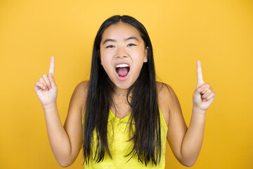 Young beautiful asian woman over yellow isolated background amazed and surprised looking at the camera and pointing up with fingers and raised arms